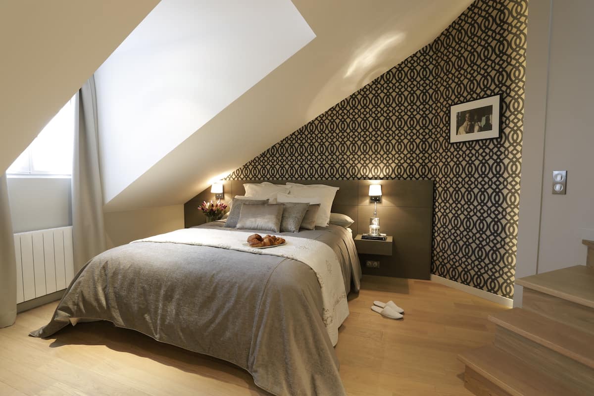 A bedroom with a sloped ceiling, a bed with a tray of pastries, bedside lamps, and a patterned wall.