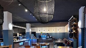 Interior modern d'un restaurant amb taules de fusta i cadires blanques, una làmpada penjant al centre i una zona de bar al fons.