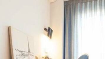 A hotel room with a bed, a desk with a chair, a painting on the wall, and a window with curtains.