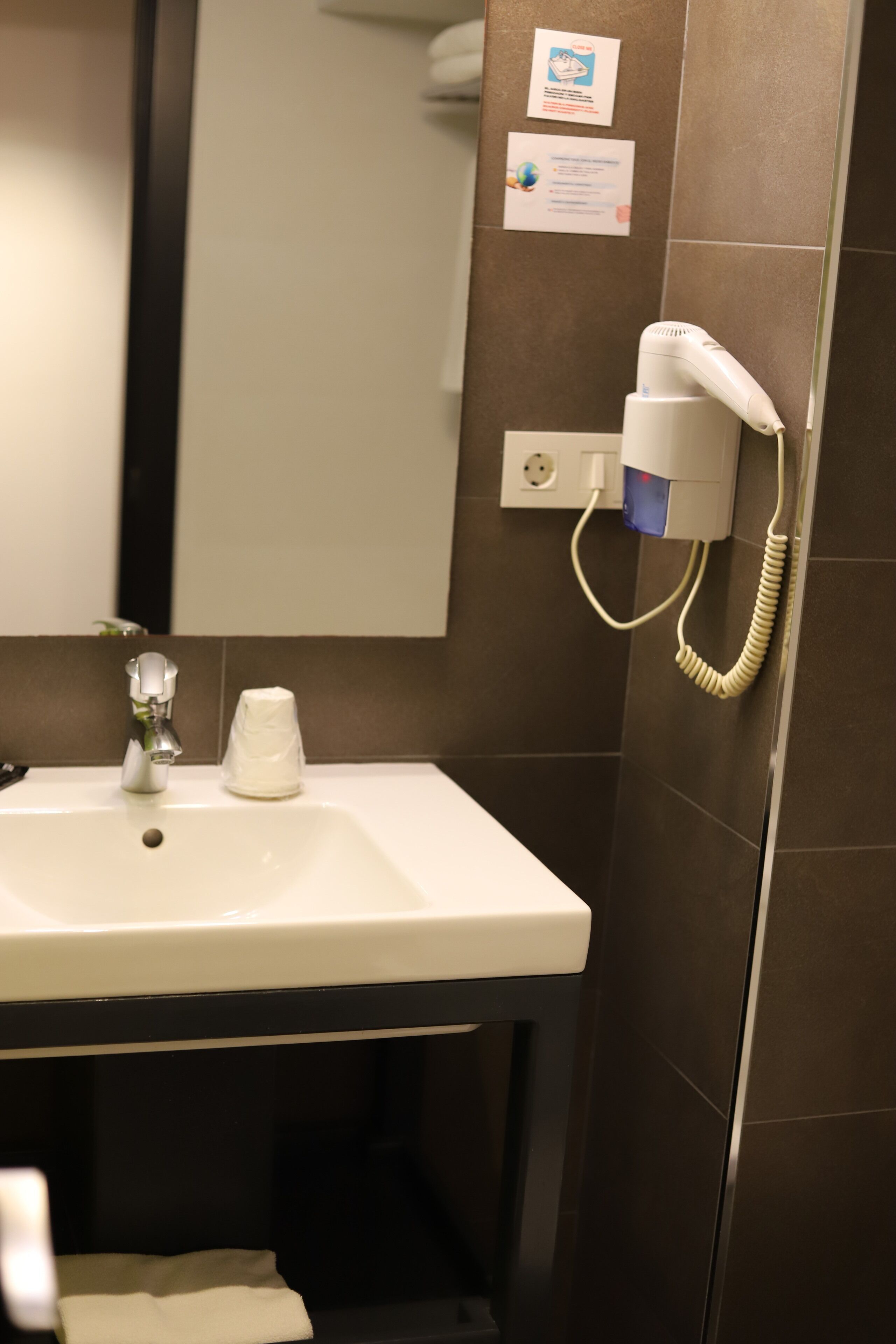 Un baño con lavamanos, espejo y secador de pelo enchufado a un enchufe.