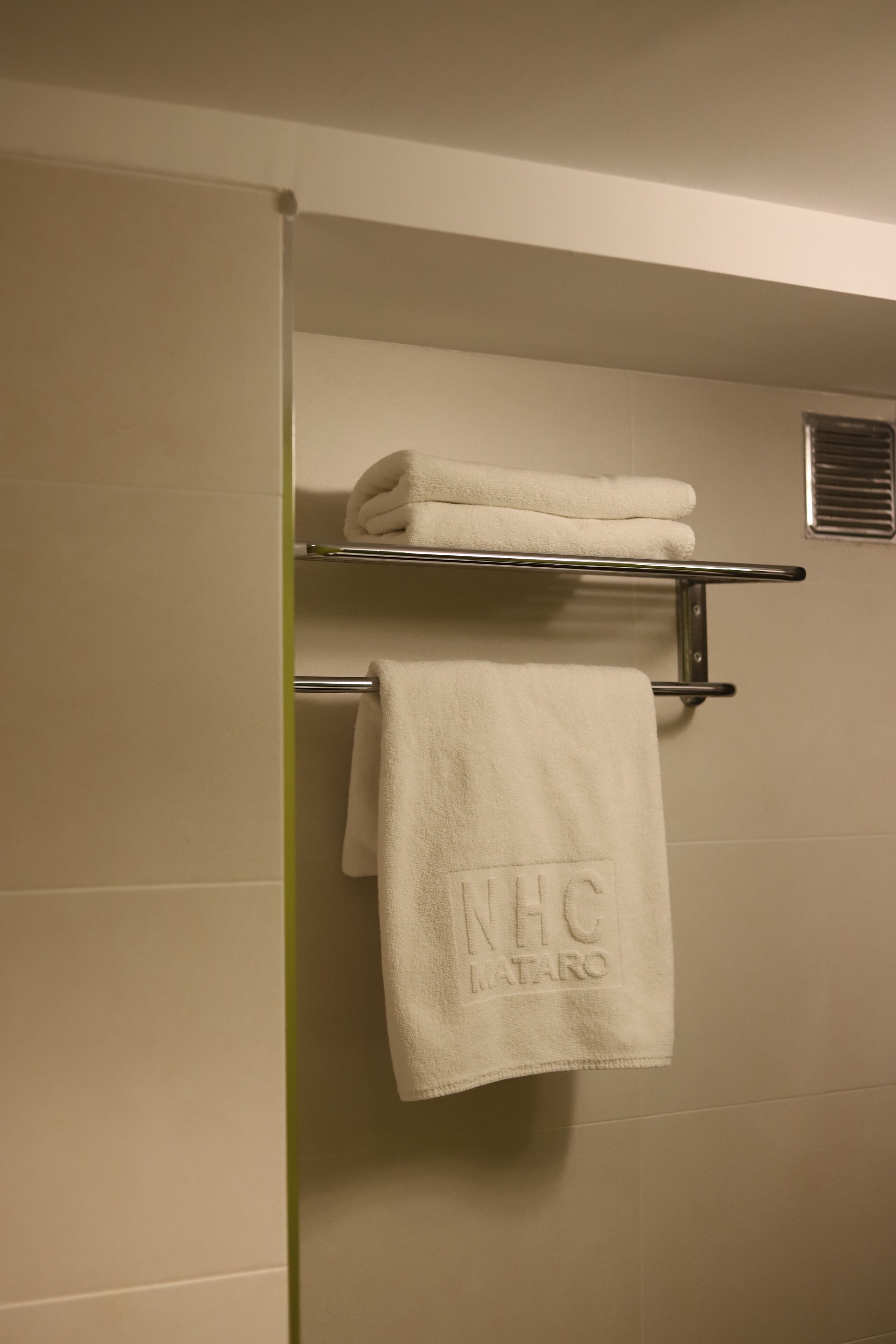 Une salle de bain avec un porte-serviettes sur lequel sont pliées deux serviettes et une autre serviette suspendue, ornée d'une broderie « NHC LATARO ».