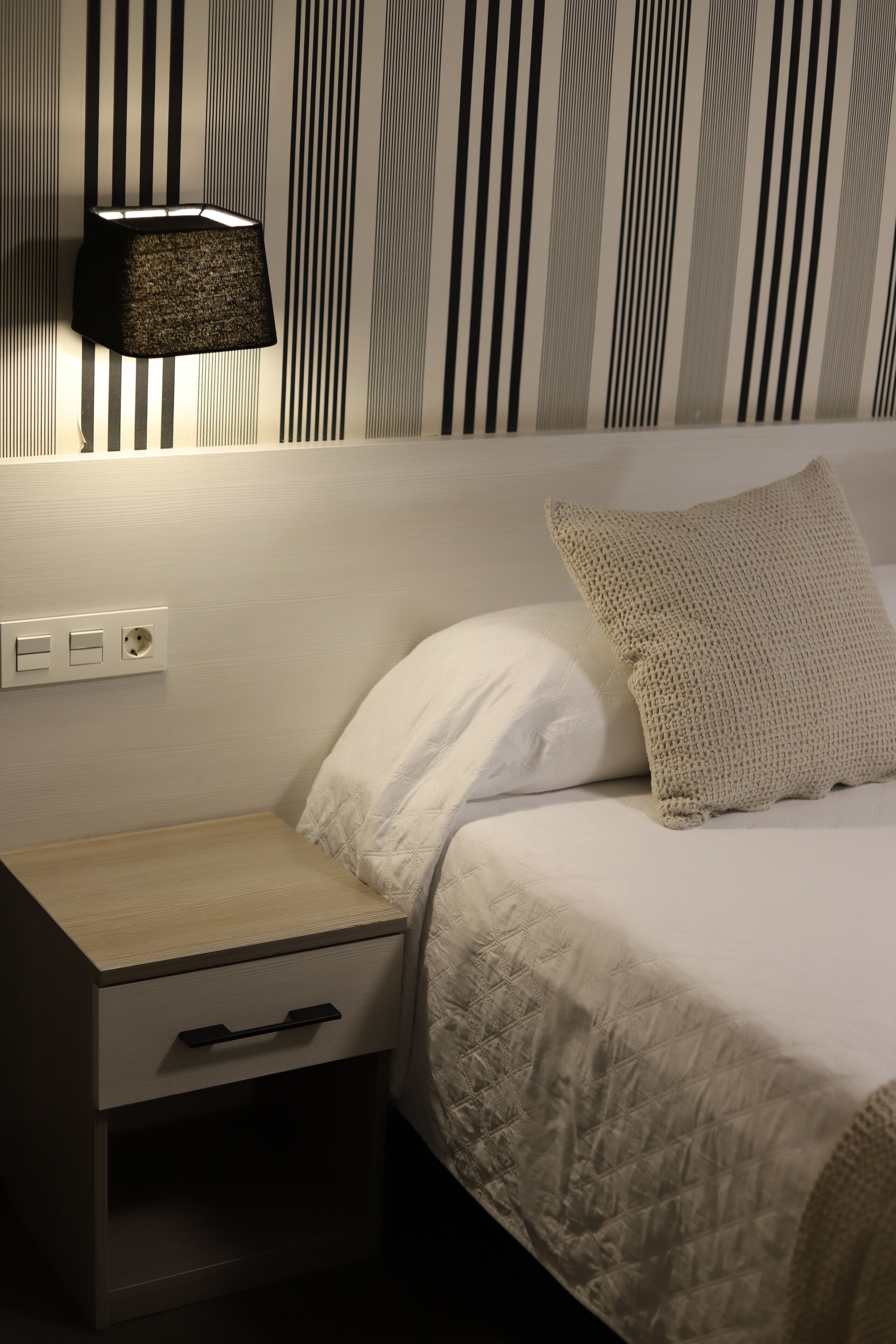 Una cama bien hecha con cubrecolcha acolchado, una lámpara de noche y una mesita de noche con cajón.