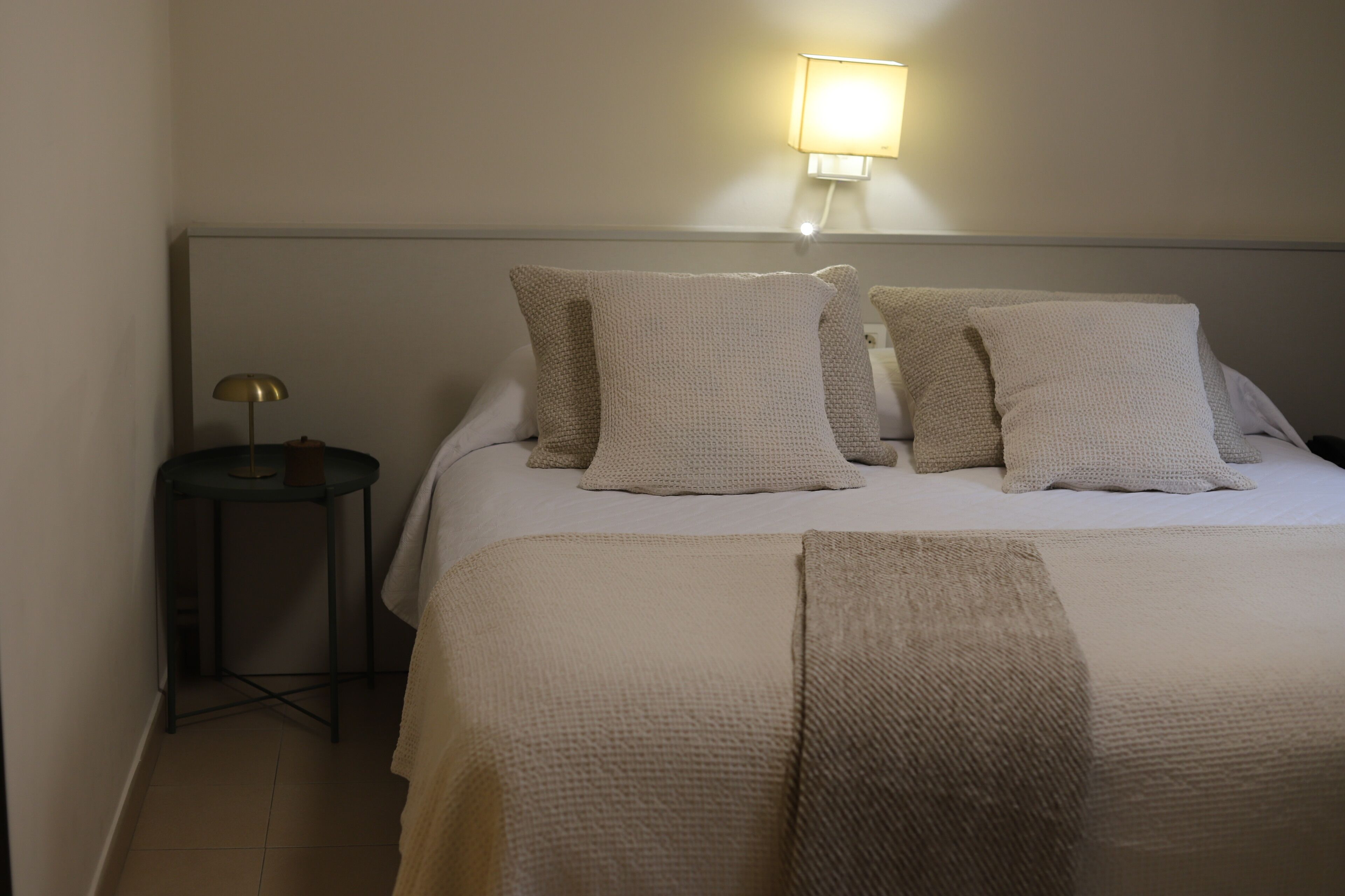 Een netjes opgemaakt bed met een grote beige deken en twee kussens, een klein rond tafeltje met een lamp, en een wandlamp.