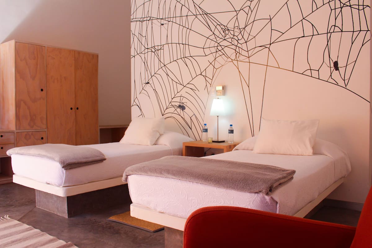 Una habitación de hotel con dos camas, un armario de madera, una obra de arte en la pared, una mesita de noche con lámpara y una silla roja.