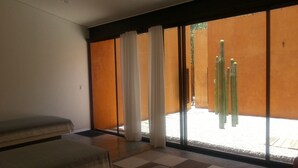 Una habitación moderna con un amplio ventanal corredero que da a un espacio exterior. Hay dos camas con ropa blanca, una mesita y un banco.