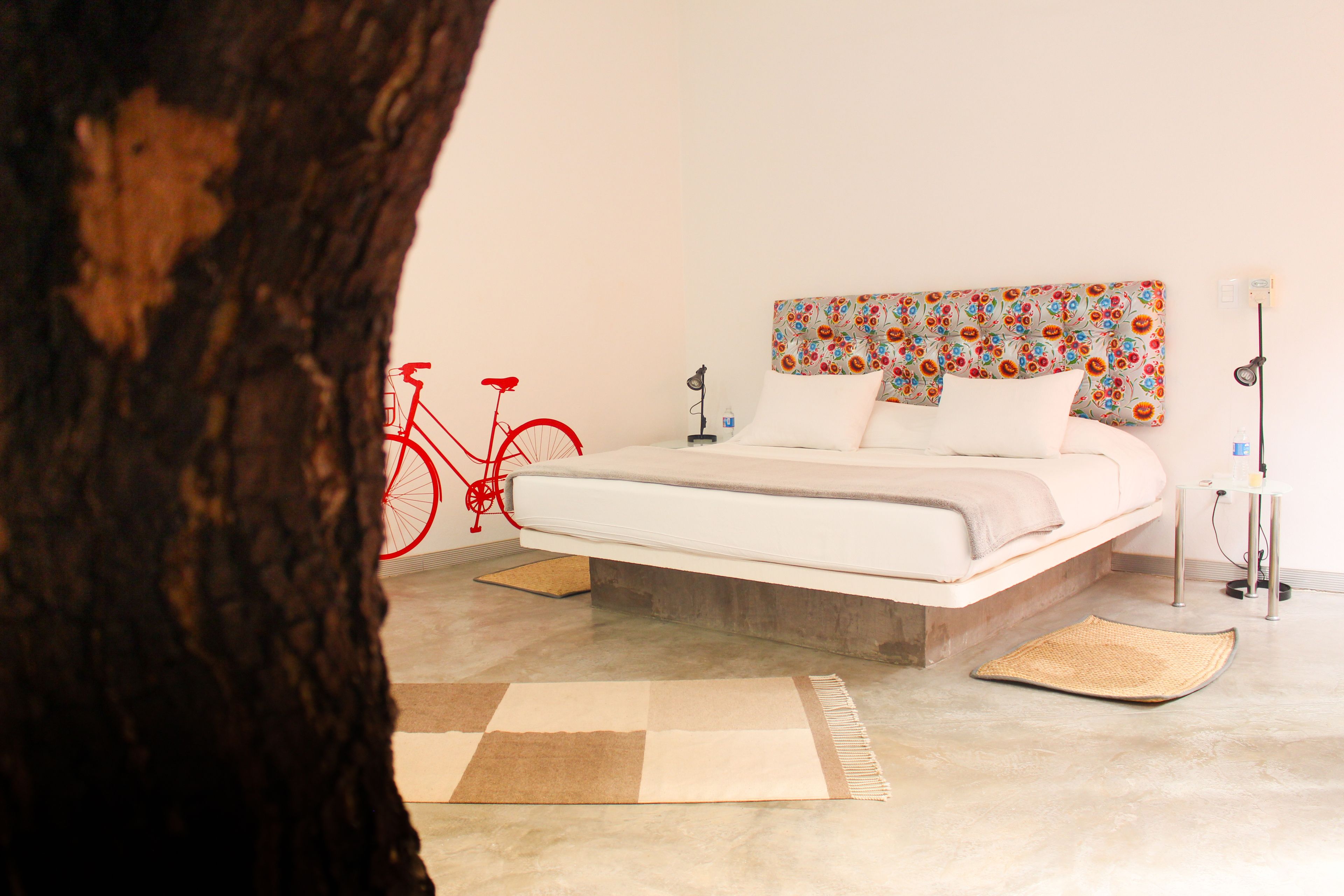 Camera da letto con un letto, una testata fiorata, una bicicletta rossa e il tronco di un albero in primo piano.