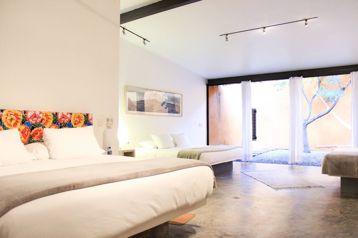 Una habitación de hotel moderna con una cama grande, una cama más pequeña y un banco. Hay una ventana con cortinas y vistas al exterior.
