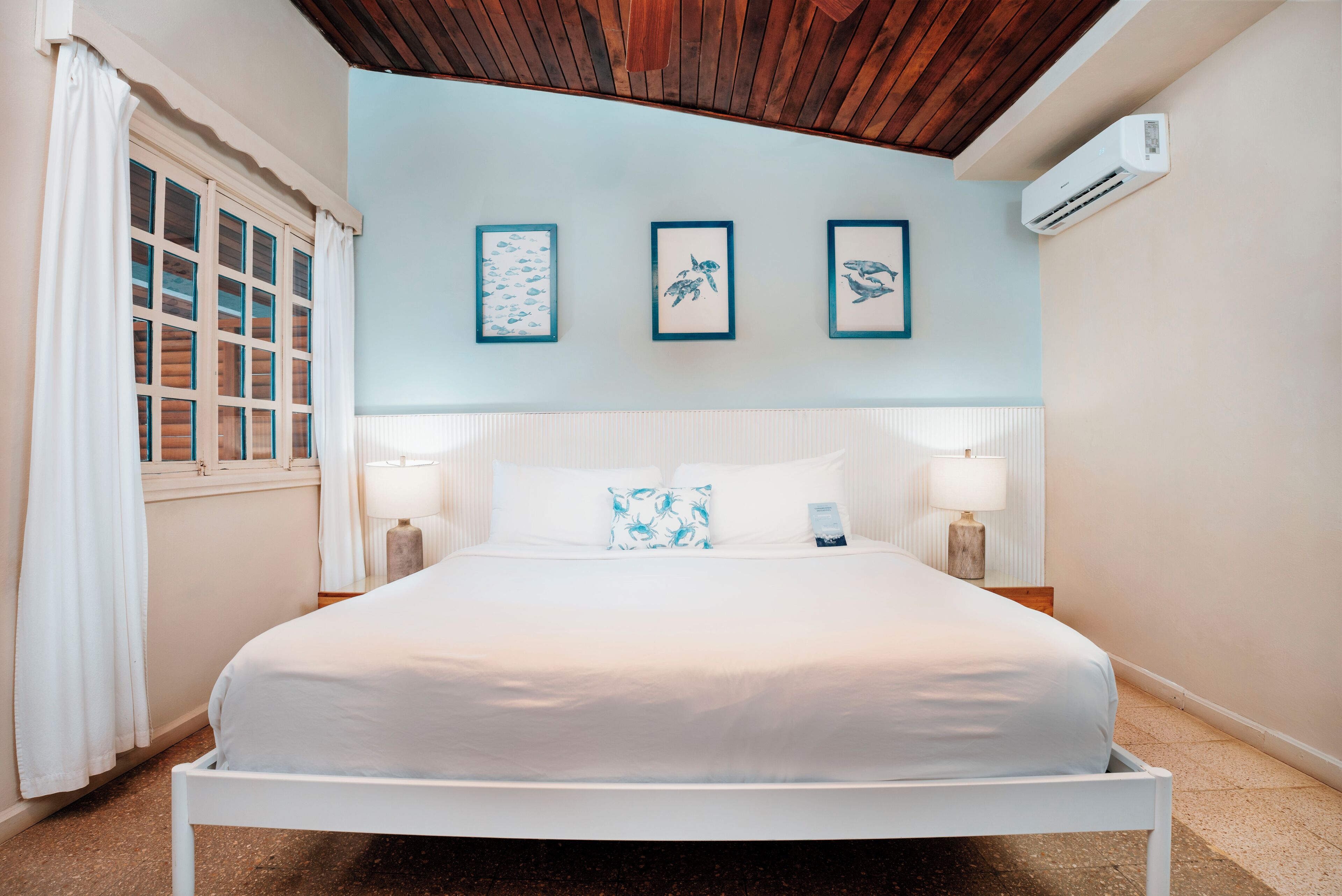 A neatly made bed with white linens, a wooden ceiling, and a wall-mounted air conditioner.