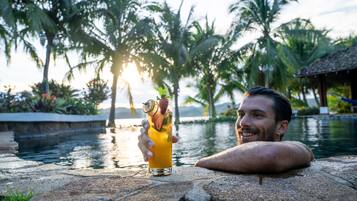 Un hombre descansando junto a una piscina con una bebida en la mano, rodeado de palmeras y un atardecer.