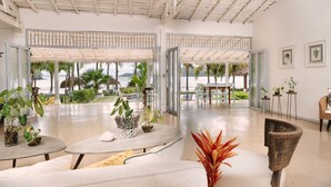Un vaste espace intérieur doté d’un haut plafond, de grandes fenêtres et d’une vue sur la plage. On y trouve plusieurs tables et chaises, et des plantes en pot sont disposées dans toute la pièce.