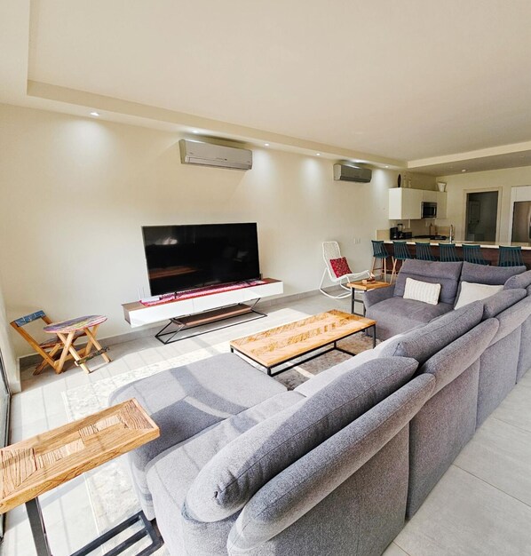 Ein modernes Wohnzimmer mit grauem Sofa, einem Holztisch, einem Flachbildfernseher und einer kleinen Essecke mit Barhocker.