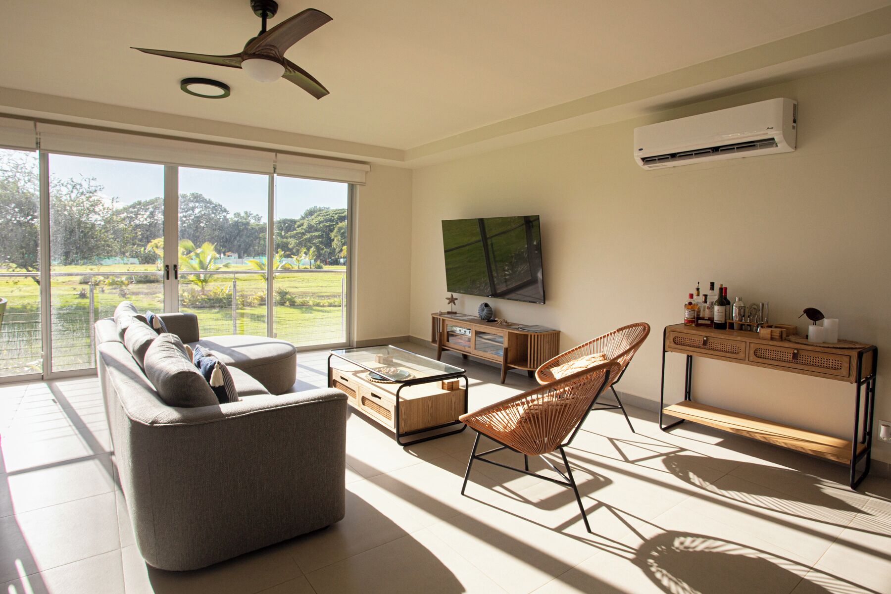 A spacious living room with a large window, a ceiling fan, a flat-screen TV, and a wooden console table with bottles.