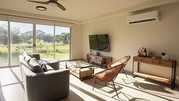 A spacious living room with a large window, a ceiling fan, a flat-screen TV, and a wooden console table with bottles.