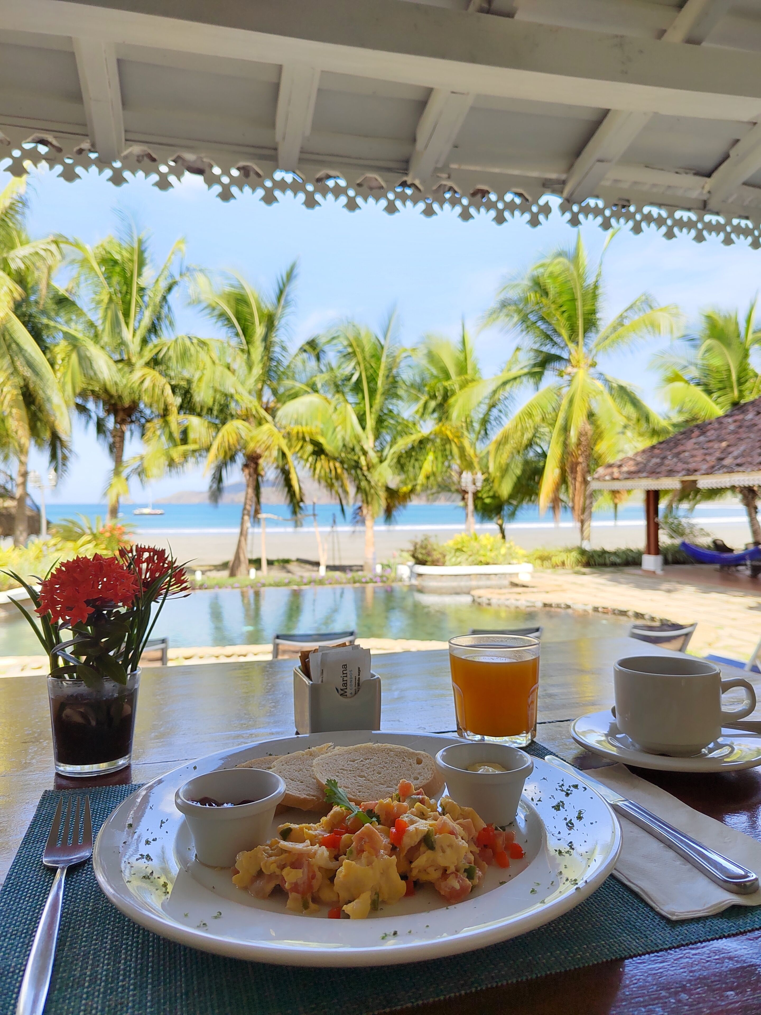 A breakfast table with scrambled eggs, toast, and a drink, overlooking a beach with palm trees and a pool.