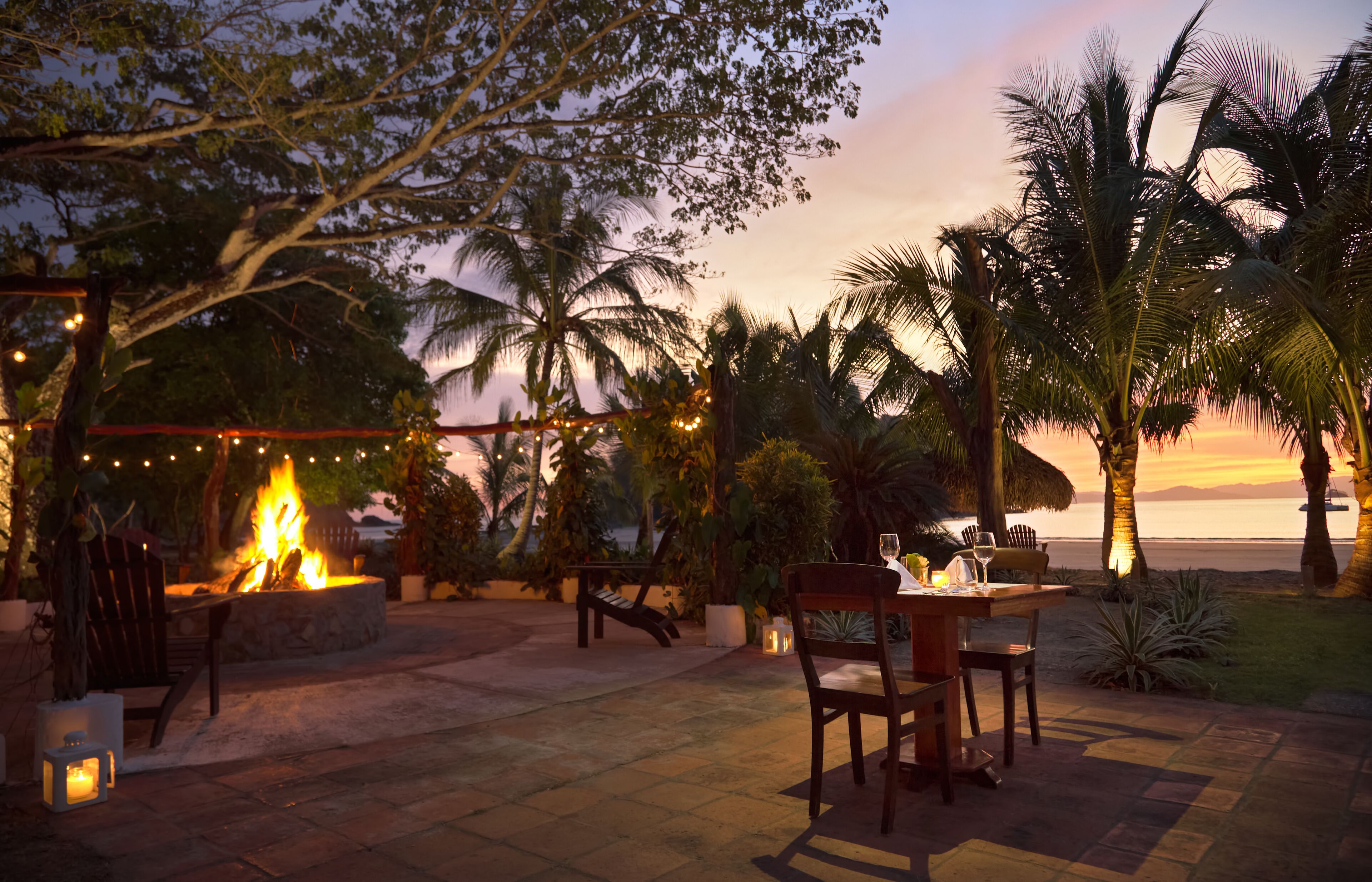 Un patio exterior con una fogata, asientos y vistas al atardecer sobre el océano.