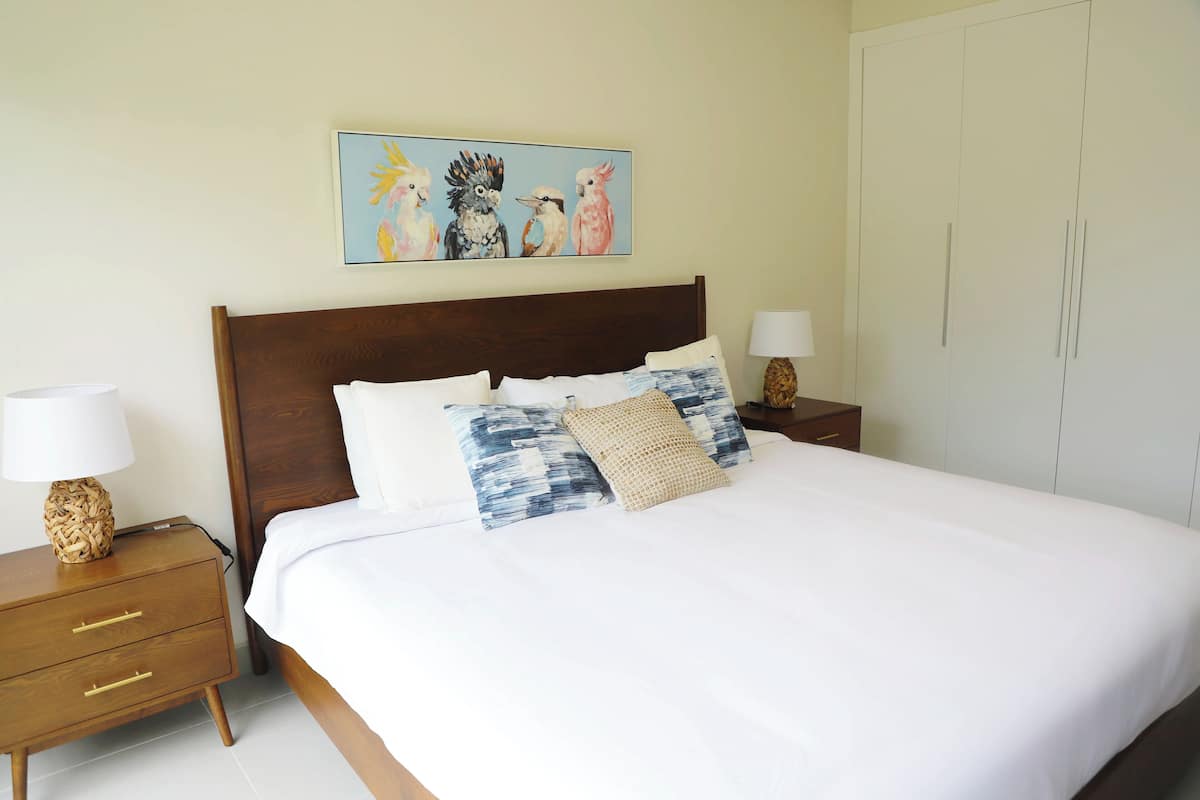 Een netjes opgemaakt bed met witte beddengoed en decoratieve kussens, een houten hoofdbord en een nachtkastje met een lamp.