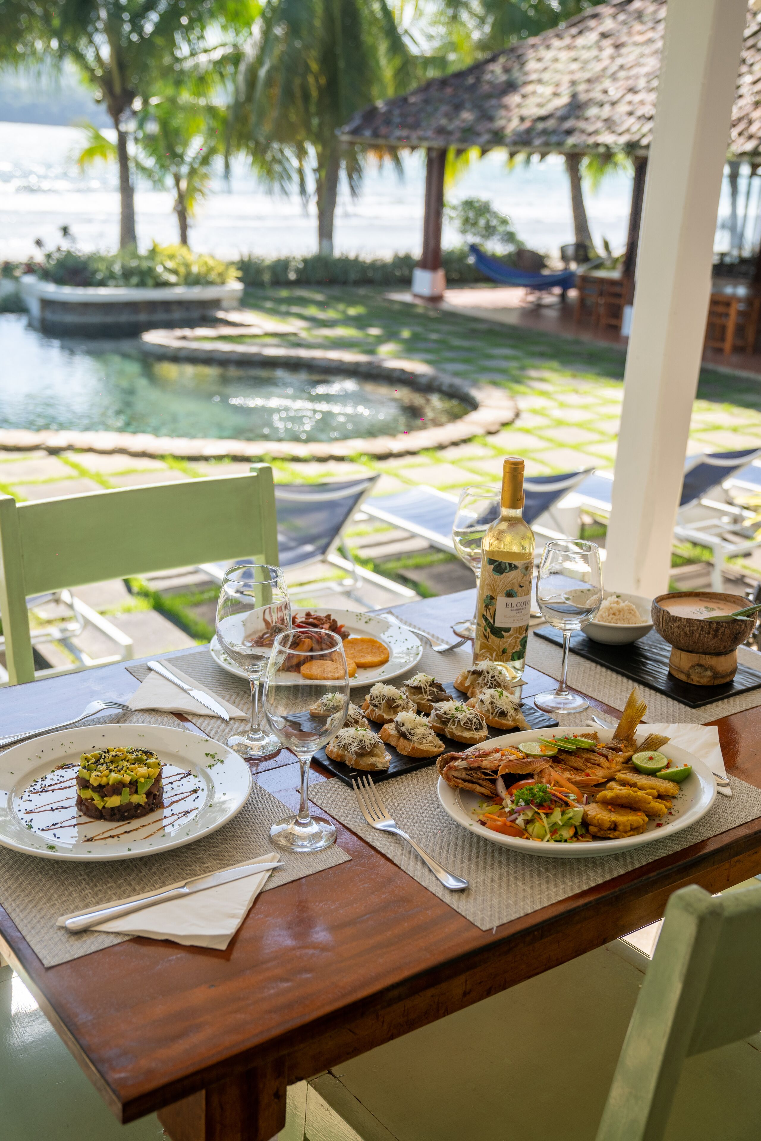Una mesa de comedor dispuesta con diversos platos, una botella de vino y vistas a una piscina y palmeras.