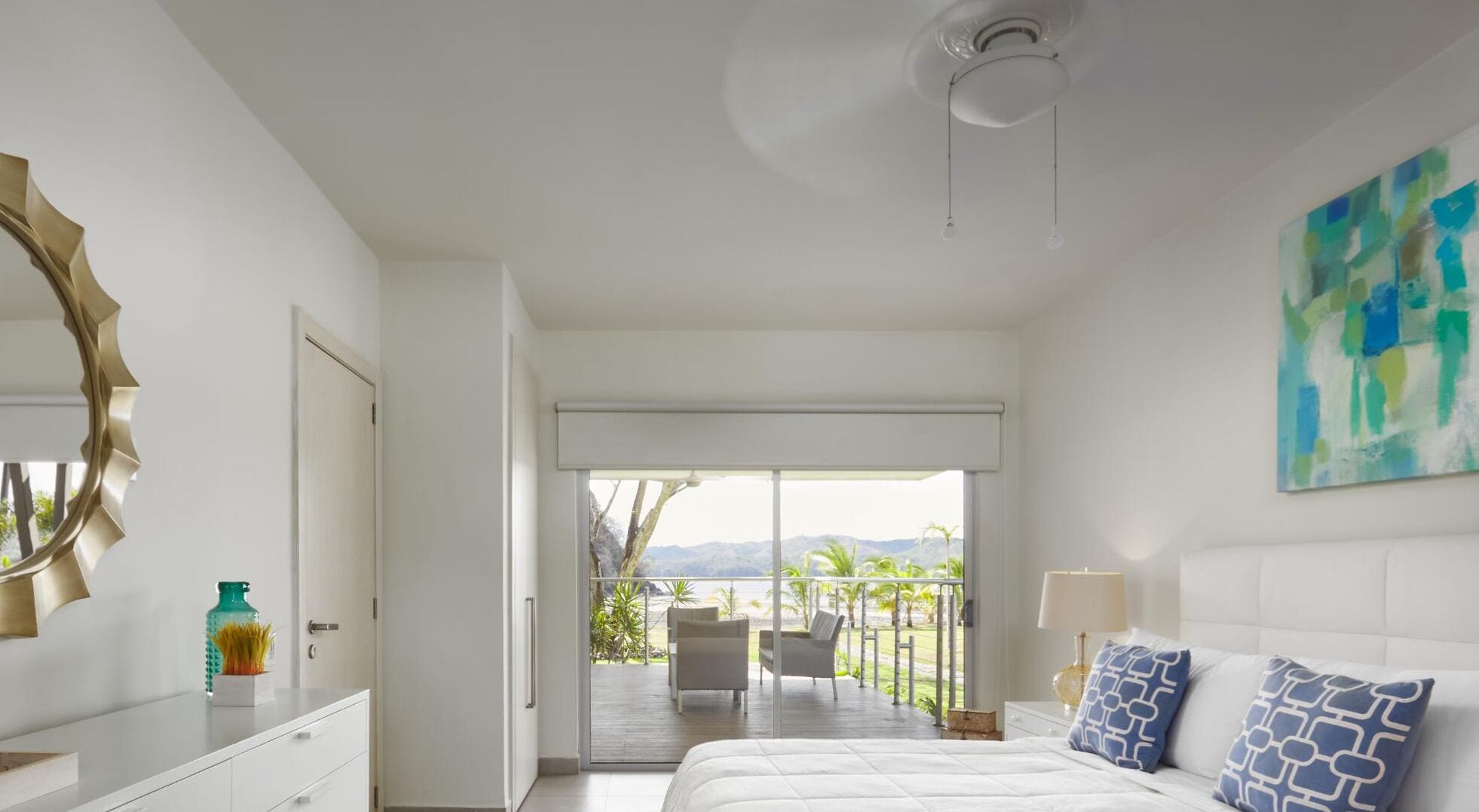 A bedroom with a large bed, a view of a balcony with outdoor furniture, and a ceiling fan.