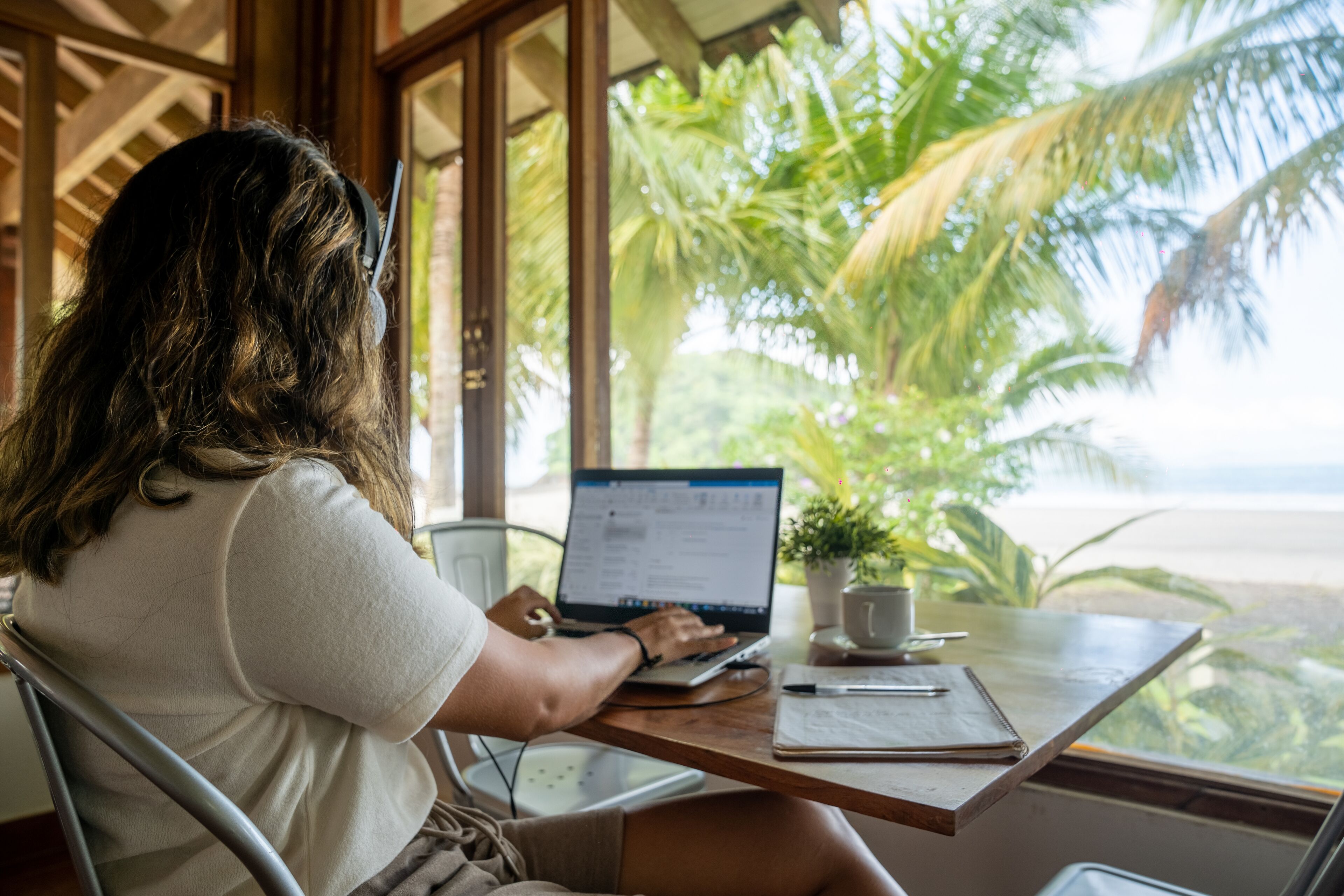 A person working on a laptop at a wooden table with a view of palm trees and the beach.