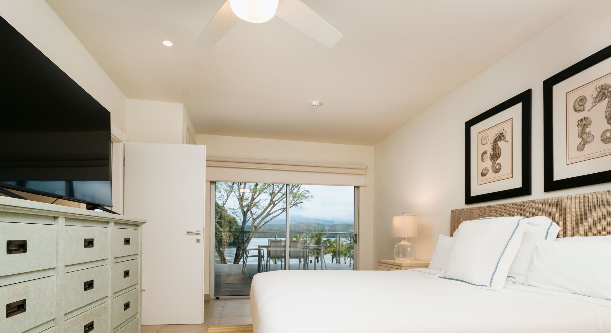 A hotel room with a bed, a television, a dresser, a balcony with a view of the water, and framed artwork on the wall.
