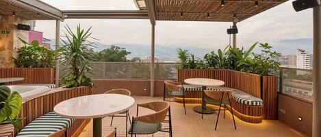 A rooftop terrace with round tables and cushioned seating, surrounded by potted plants and a city view.