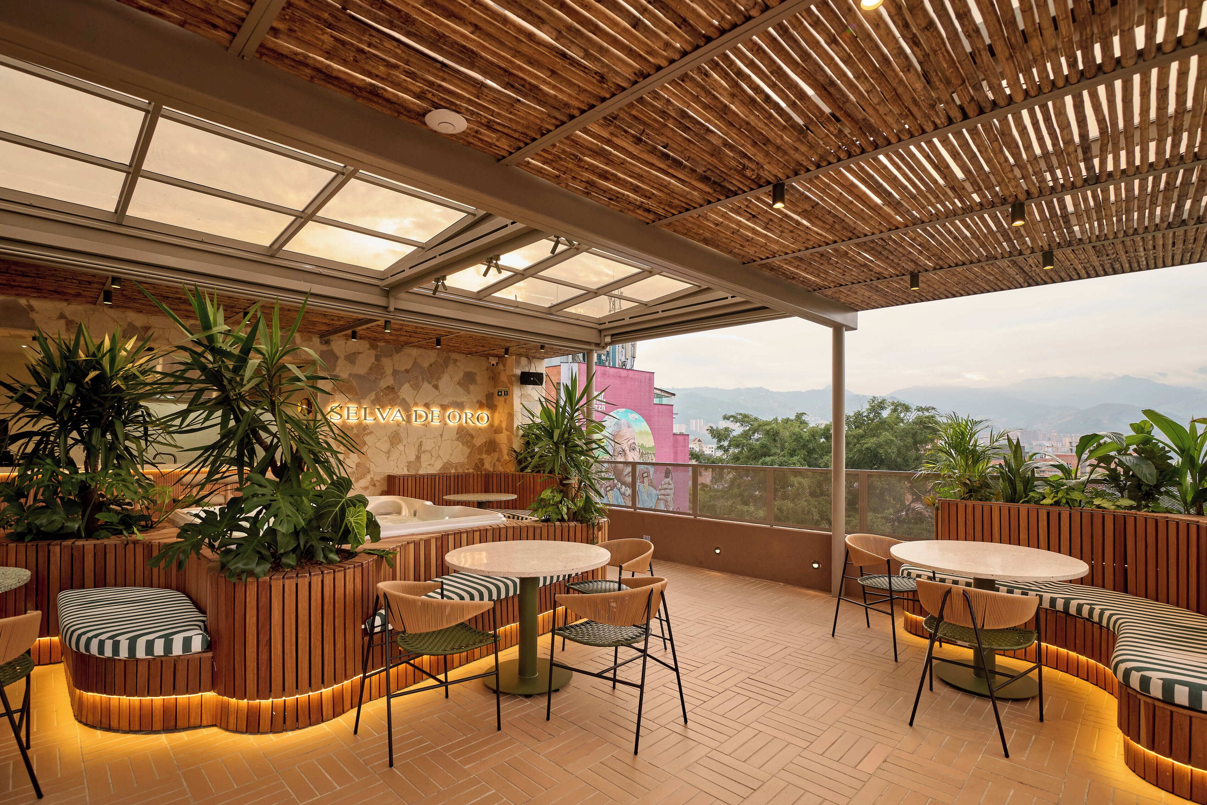 A rooftop terrace with wooden flooring, round tables, and chairs, surrounded by potted plants and a mural.