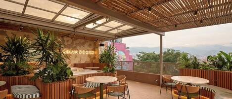 A rooftop terrace with wooden flooring, round tables, and chairs, surrounded by potted plants and a mural.