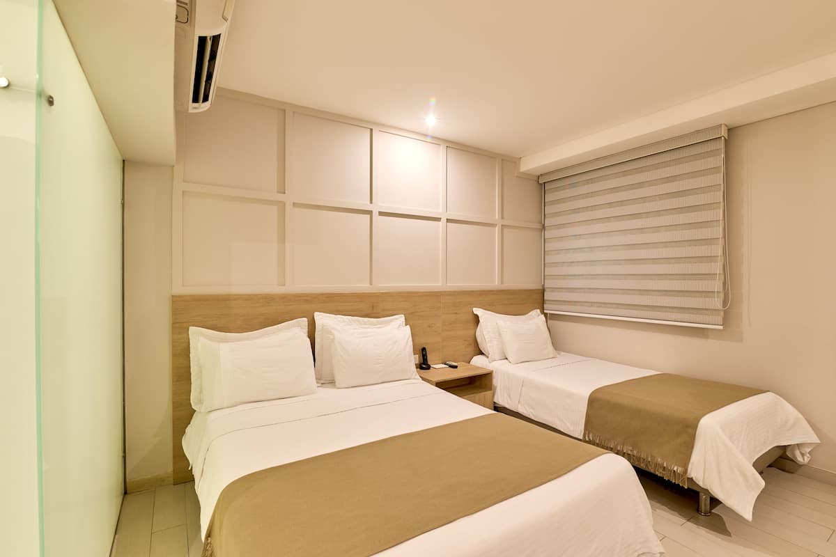 Une chambre d’hôtel avec deux lits, une tête de lit en bois, une table de chevet, un téléphone et une fenêtre avec des stores.