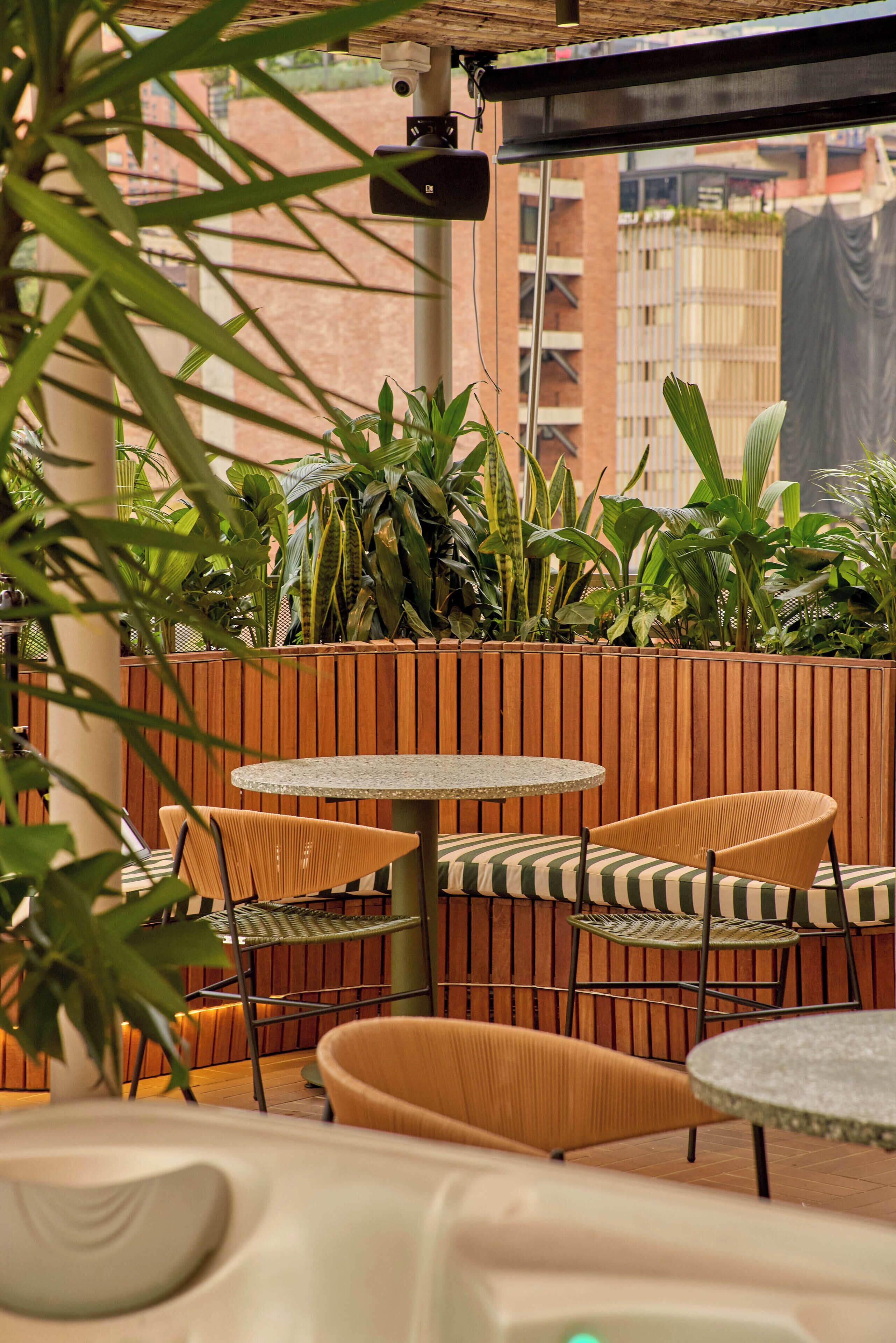 An outdoor seating area with round tables and wicker chairs, surrounded by a wooden fence and lush greenery.