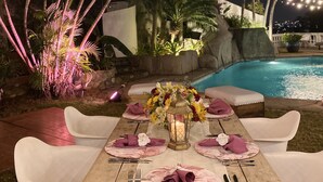 Un área de comedor al aire libre con una mesa de madera dispuesta para cuatro personas, rodeada de sillas blancas, bajo luces colgantes.