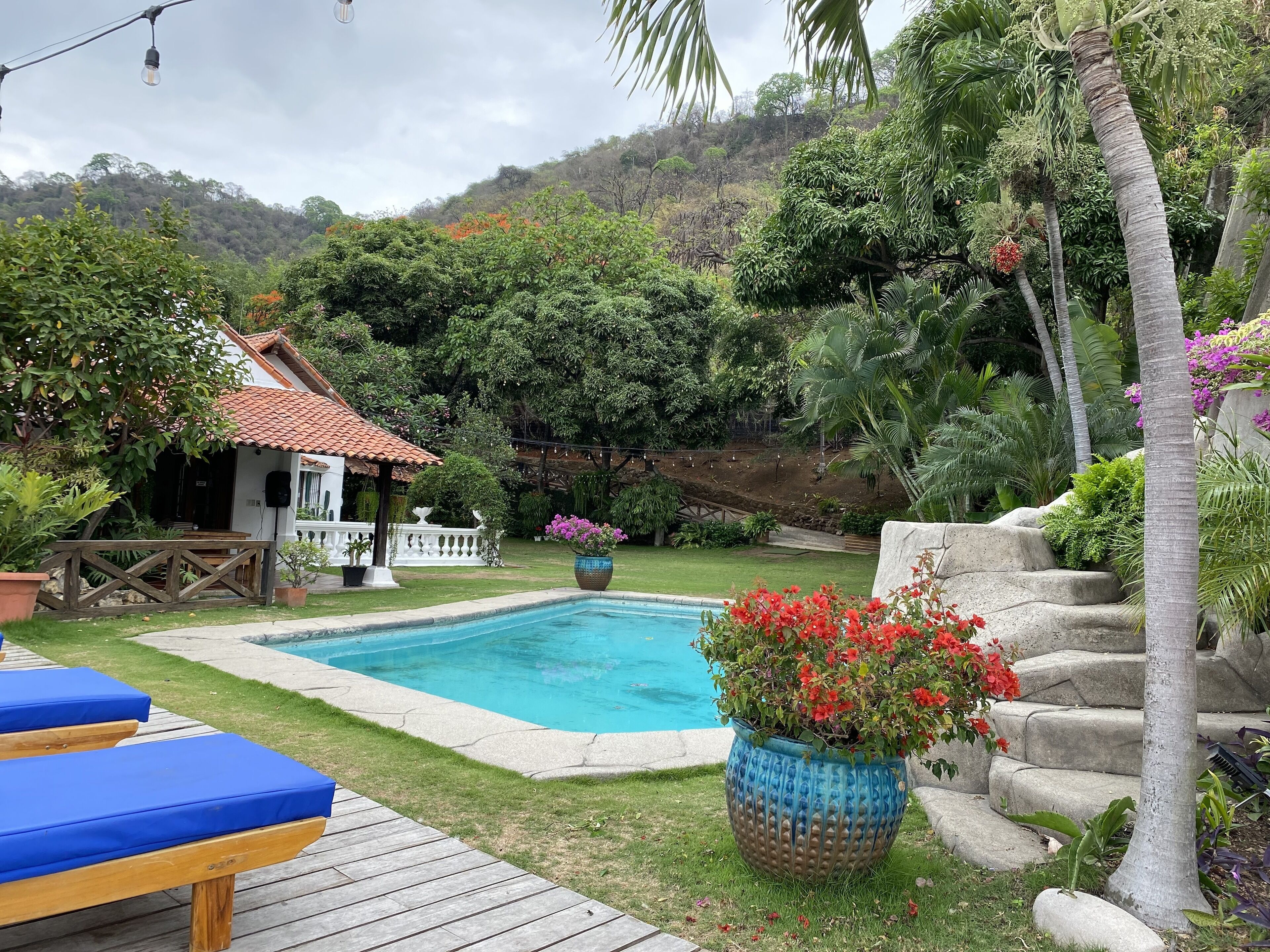 Un área de piscina con terraza de madera, sillas de descanso, un pequeño edificio con techo de tejas y exuberante vegetación alrededor de la propiedad.