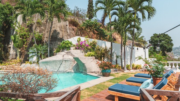 Un área de piscina con sillones de descanso, una cascada y plantas tropicales.