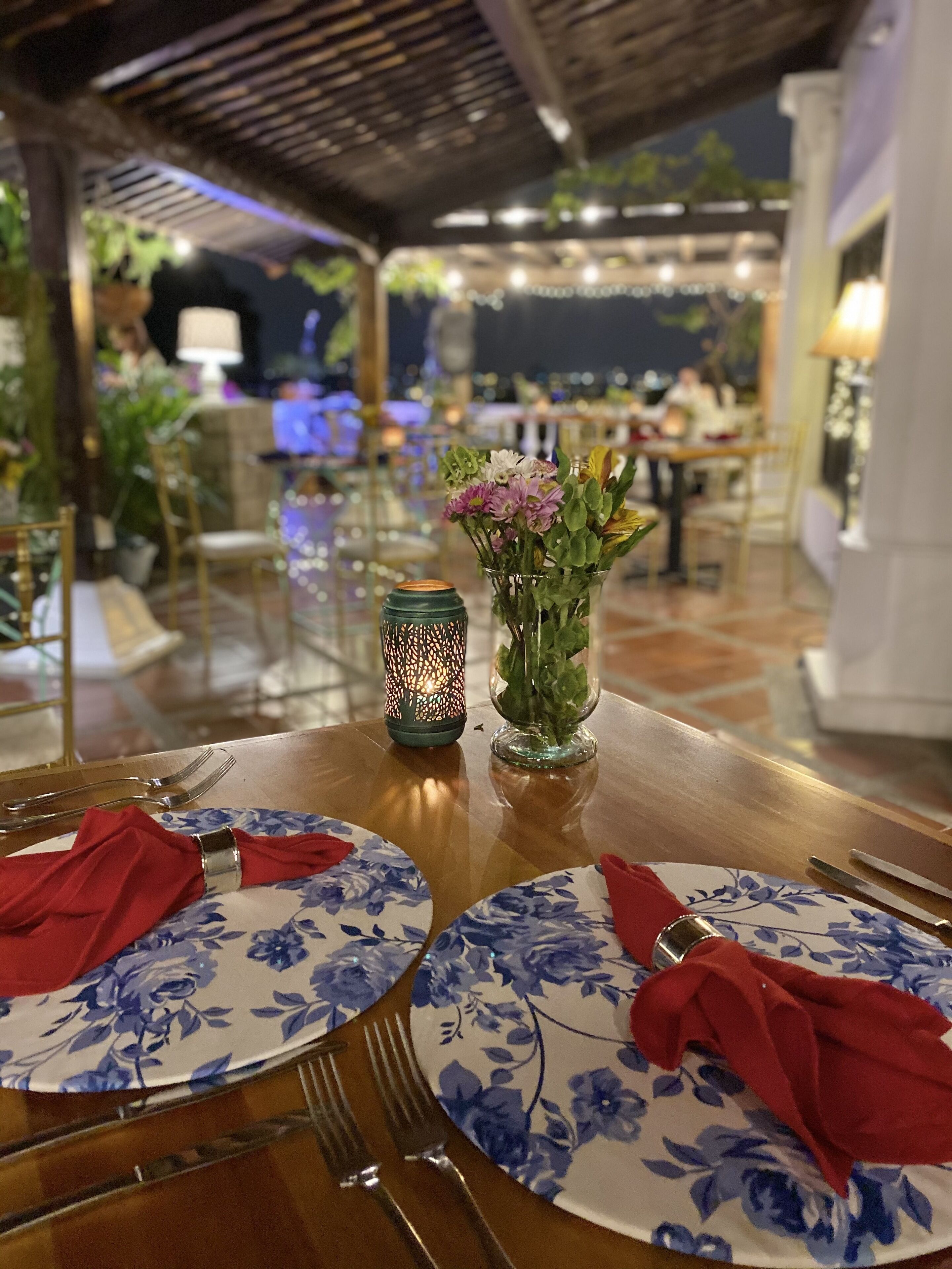 Una mesa de comedor vestida con manteles florales, servilletas rojas y un jarrón decorativo con flores.