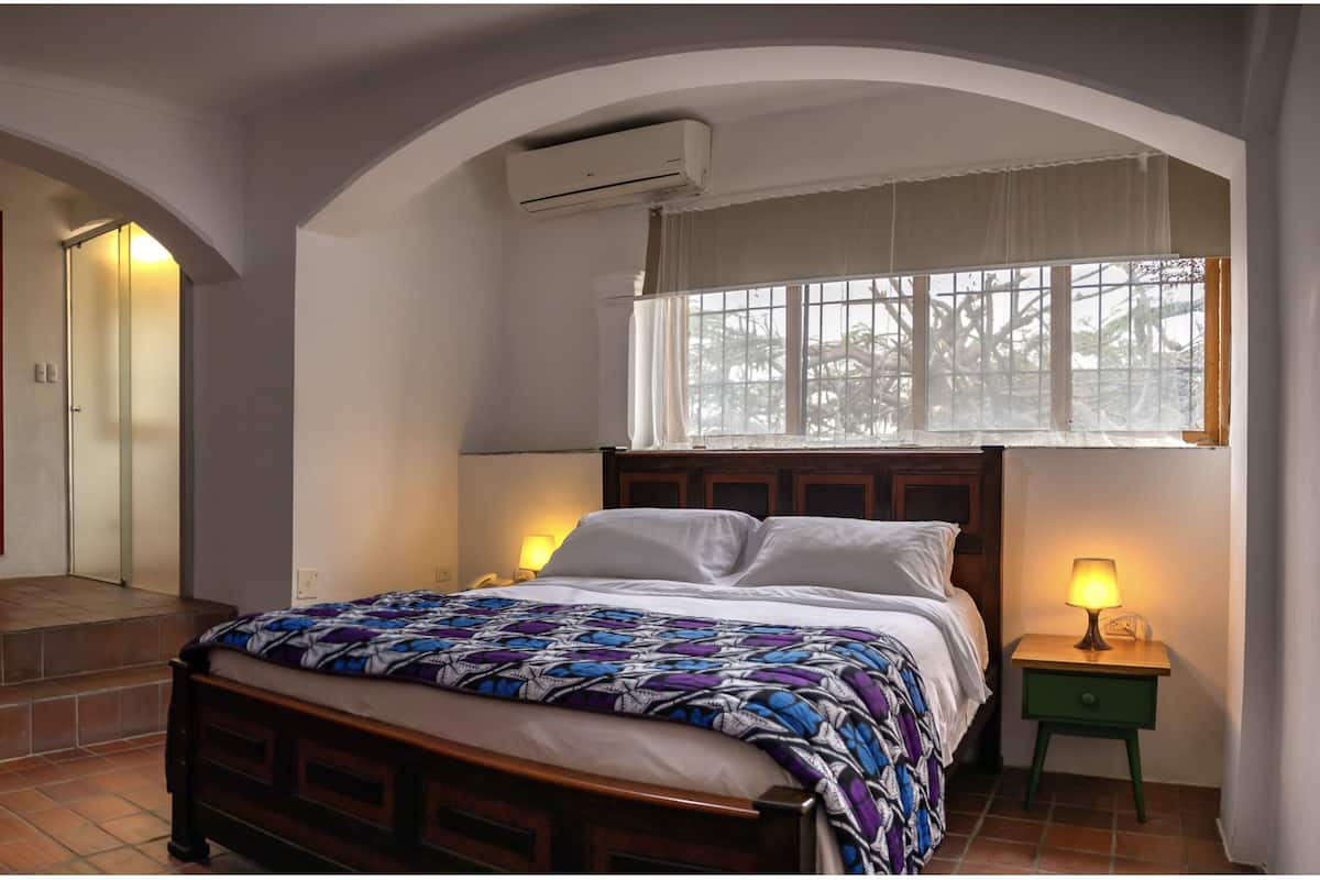 Un dormitorio con una cama, una mesita de noche, una lámpara, una puerta roja, una escalera y un ventanal de arco.