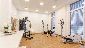 A modern fitness room with various exercise equipment, a wooden floor, and a large window with blinds. - PLAZA INN Dachau - München (Dachau)