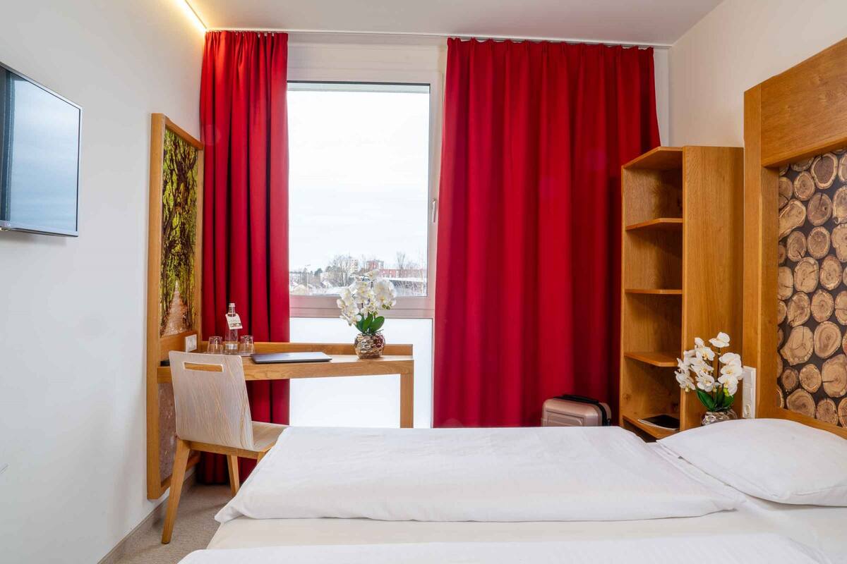 A hotel room with a bed, a desk with a chair, a TV, and a large window with red curtains.