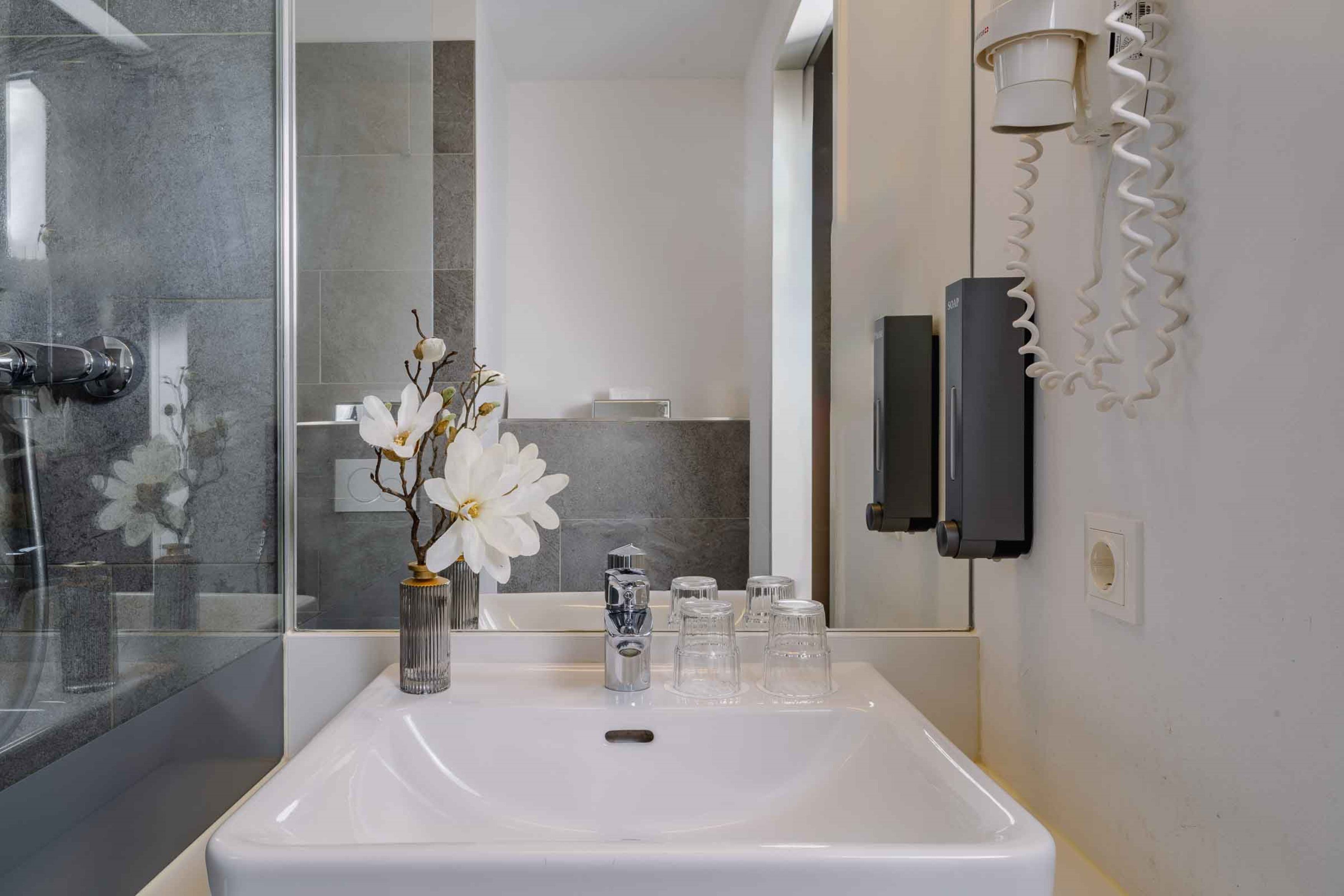 Un baño moderno con un lavamanos blanco, una cabina de ducha con paredes de vidrio y un florero con flores blancas.