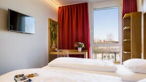 A hotel room with a bed, a desk, a TV, and a window with red curtains. - PLAZA INN Dachau - München (Dachau)