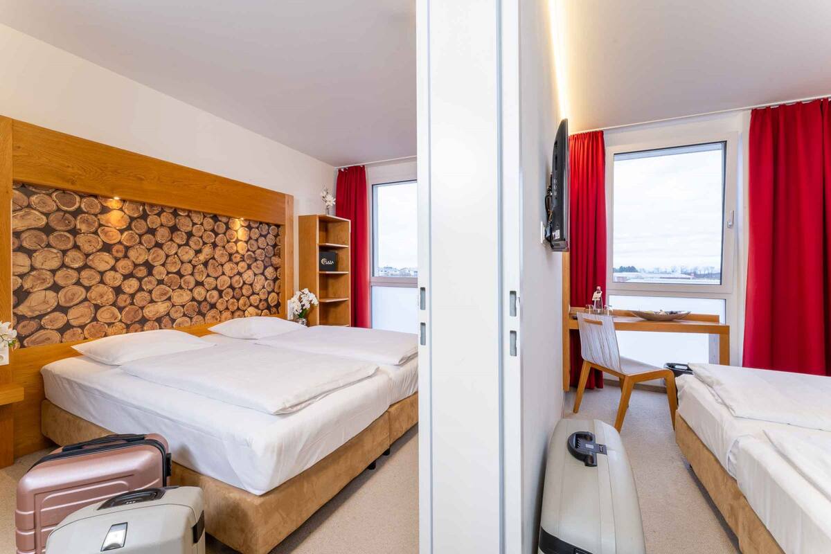 A hotel room with two beds, a wooden headboard, a desk with a chair, a TV, and a window with red curtains.