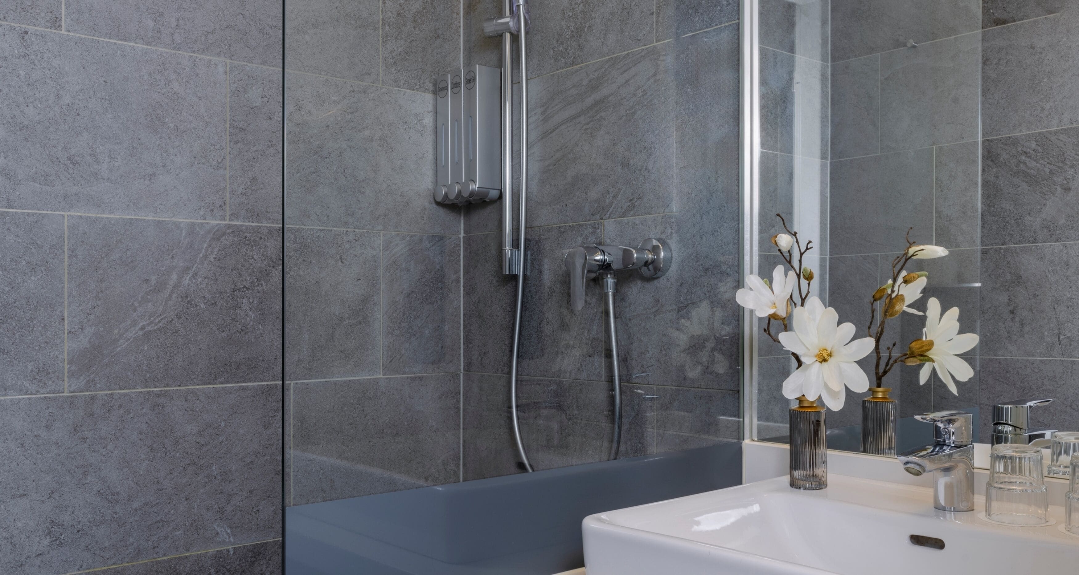 Een moderne badkamer met grijze tegels, een badkuip, een douche en een wastafel met een vaas witte bloemen.