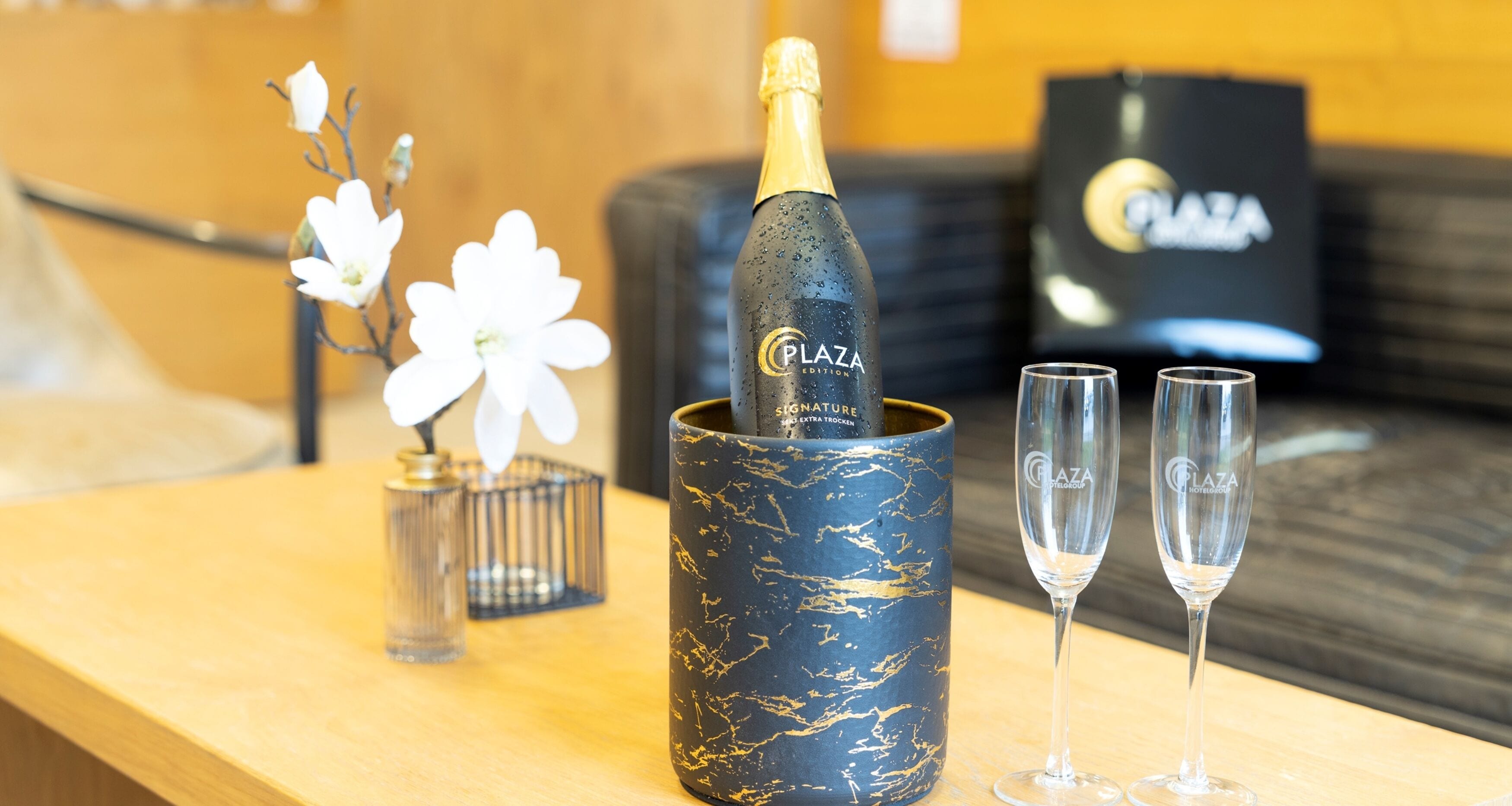 A bottle of champagne in a decorative holder with two glasses beside it, set on a wooden table with a vase of flowers and a black bag in the background.