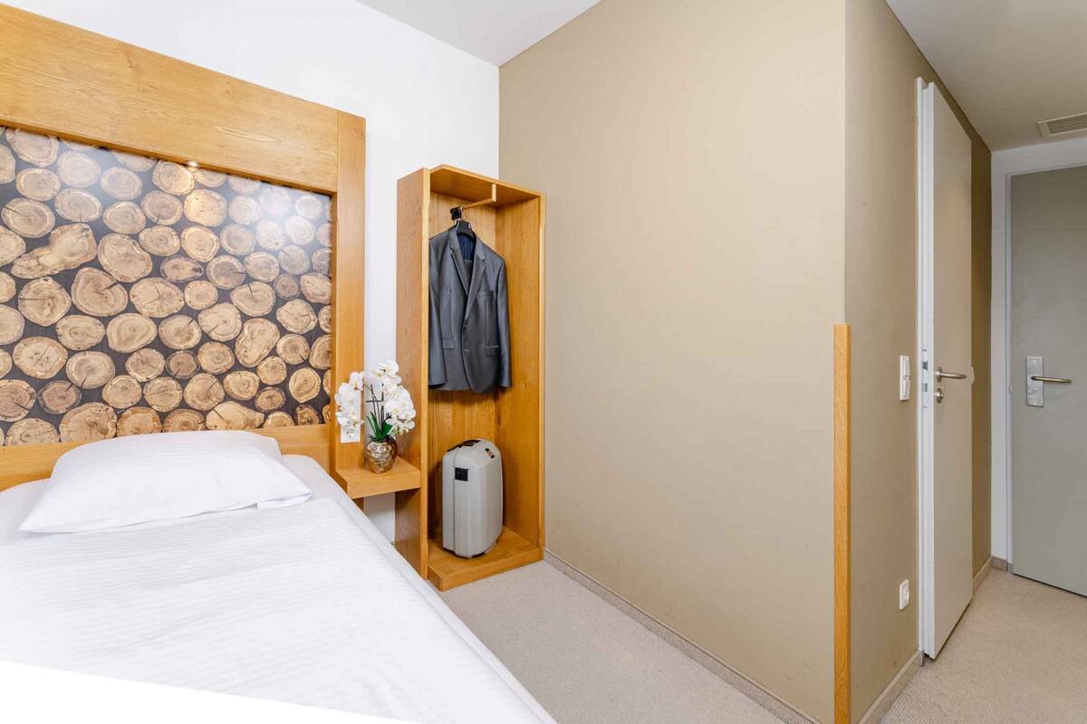 A hotel room with a bed, a wooden headboard, a small shelf with a vase of flowers, a coat hanging, and a door leading to another room.