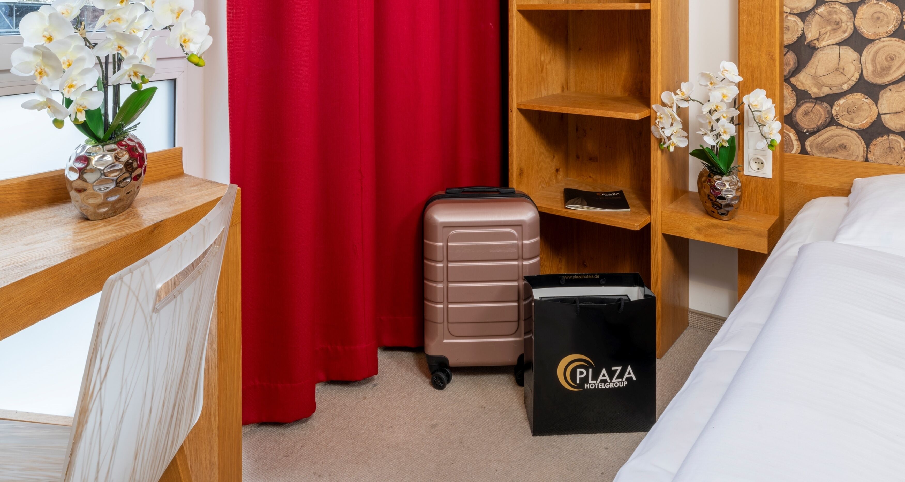 A hotel room with a bed, a wooden desk, a red curtain, a suitcase, and a black shopping bag with the logo "Plaza Hotel Group".