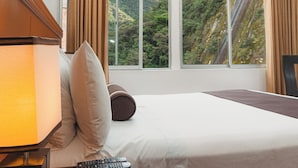 A hotel room with a bed, a lamp, a remote control, and a window with a view of a forested hillside. - Hotel Ferre Machu Picchu (Machu Picchu)
