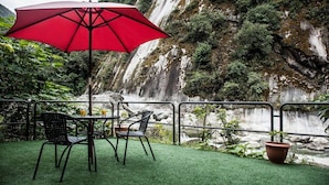 An outdoor seating area with a red umbrella, chairs, and a table, overlooking a waterfall and lush greenery. - Hotel Ferre Machu Picchu (Machu Picchu)