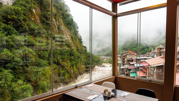 A dining area with a table set for two, large windows offering a view of a mountainous landscape and a village below. - Hotel Ferre Machu Picchu (Machu Picchu)