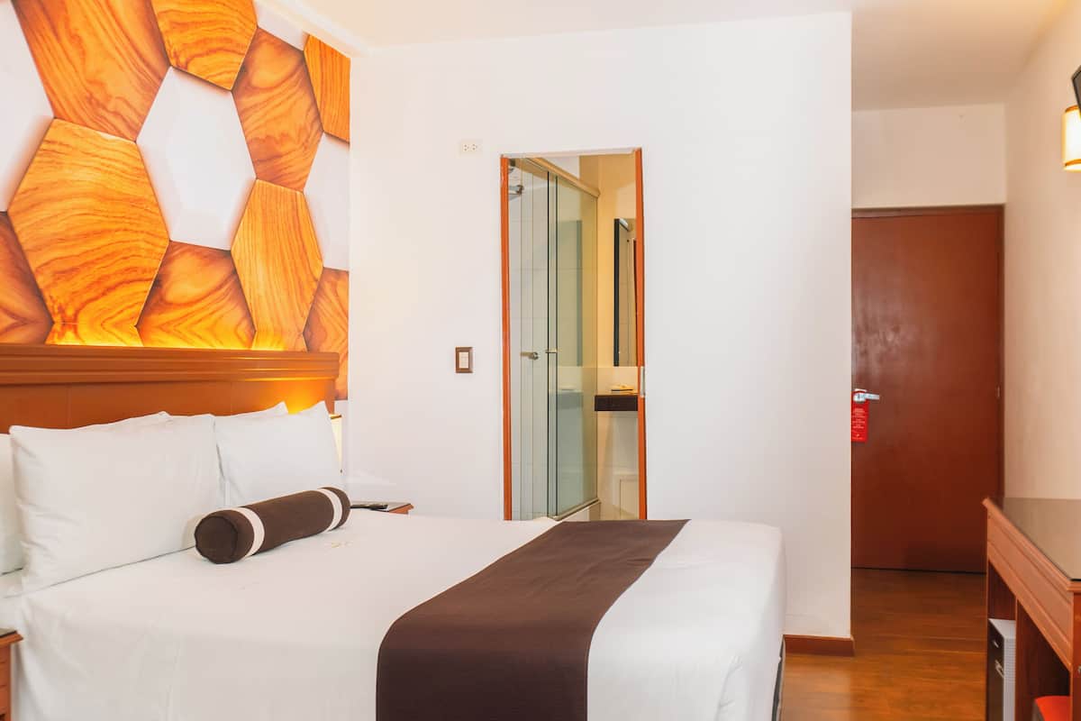 A hotel room with a bed, a wooden headboard, a glass door leading to a bathroom, and a wooden dresser.