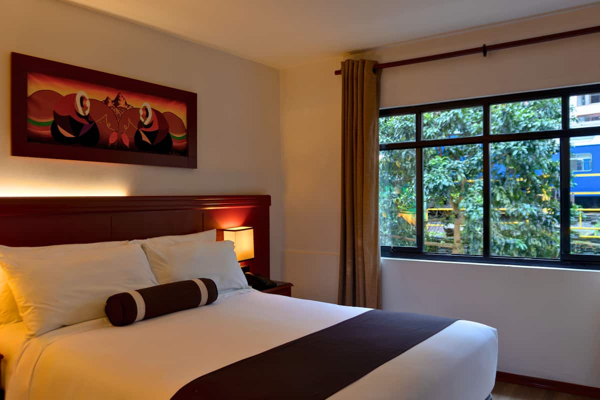 A hotel room with a large bed, a nightstand with a lamp, a painting on the wall, and a window with a view of greenery and a building outside.