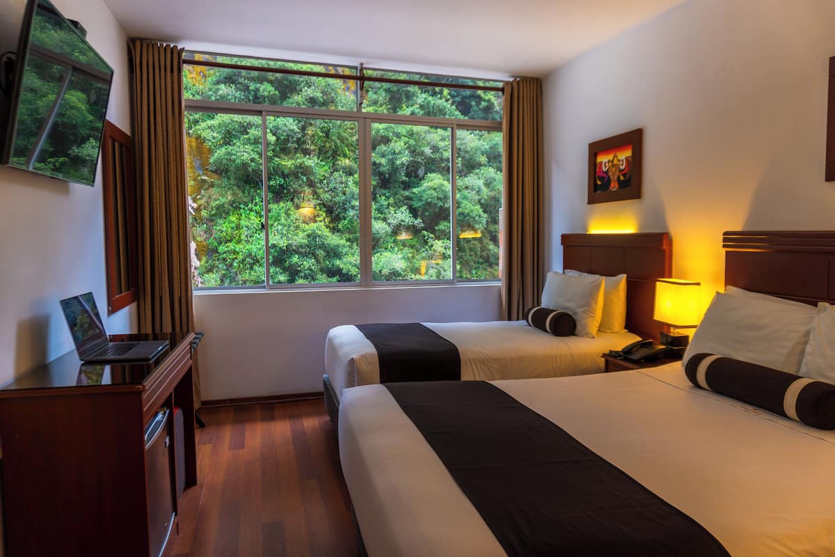 A hotel room with two beds, a desk with a laptop, a nightstand with a lamp, and a large window with a view of trees.