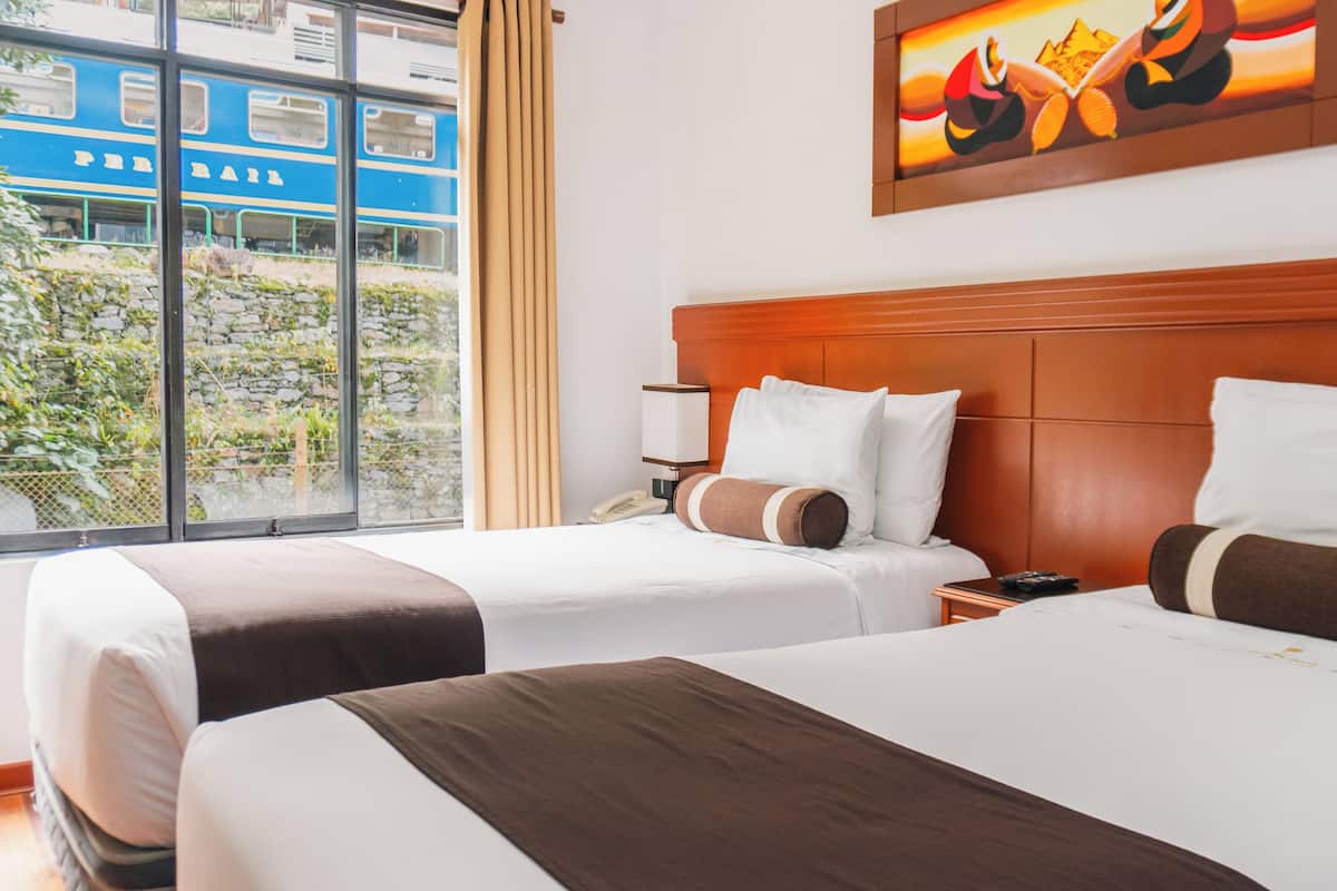 A hotel room with two beds, a wooden headboard, a bedside table with a lamp, and a window with a view of a train.